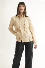 Camisa color crema con cuello clásico, cierre frontal con botones y mangas largas. Presenta detalles de bordado calado en el frente y los puños.