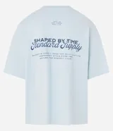 Remera de algodón celeste claro, corte oversized, con cuello redondo y manga corta. Presenta un pequeño texto bordado en azul oscuro en el pecho a cada lado.