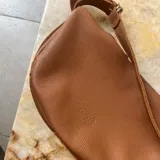 Cartera bandolera de cuero graneado color marrón, con correa ajustable que rodea toda la cartera. Tiene forma de media luna con bordes redondeados y logo Loria Studio grabado en el frente. Interior forrado en algodón.