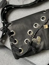 Cartera de cuero negra con correa regulable, estilo bandolera o al hombro. Decorada con charm de corazón metálico, encaje negro, cadenas y aros metálicos.