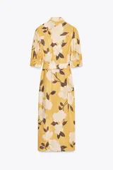 Vestido midi camisero color amarillo con estampado de flores blancas y hojas marrones. Tiene cuello solapa, mangas 3/4 con puños, cinturón de lazo ajustable y cierre frontal con botones.