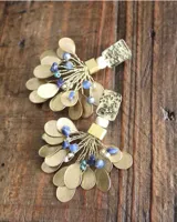 Par de aros colgantes con base rectangular dorada texturada, cuentas de color blanco y dorado, y colgantes de metal dorado en forma de hojas y cuentas de color azul.