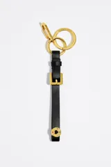 Llavero con correa de piel negra que simula una hebilla dorada, mosquetón y anillas doradas. Incluye un dije circular dorado con un corazón esmaltado en colores.