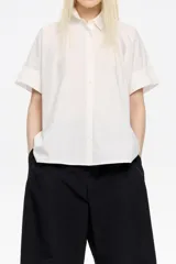 Camisa blanca de corte oversize confeccionada en 100% algodón, con cuello camisero, manga corta recta y ancha, cierre frontal con botones y pespuntes a contraste.