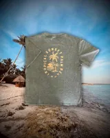 Remera de manga corta, color verde musgo con efecto gastado (acid wash). Estampado en la espalda con un diseño de atardecer tropical con palmeras y texto circular que dice "SUNSET", "TROPICAL" y "PALM BEACH".