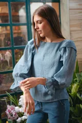 Blusa celeste de algodón con mangas largas abullonadas y puños elastizados con volados. Presenta volados en los hombros y escote redondo.
