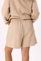 Conjunto deportivo color marrón, compuesto por un buzo con capucha, cierre frontal, bolsillos y terminaciones metálicas, y un short con cintura elástica ajustable con cordón.