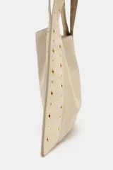 Bolso bandolera color beige de piel serraje, con tachuelas metálicas doradas en el cuerpo y las asas. Tiene doble asa de hombro, bolsillo interior y cierre con botón imantado.