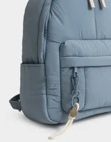 Mochila color crema de tela acolchada con pespuntes verticales. Tiene un bolsillo frontal con cierre, doble cierre superior, manija superior, correas regulables y bolsillos laterales. Incluye un charm colgante de corazón.