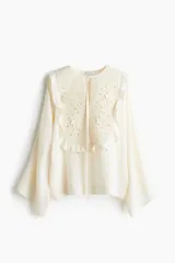 Blusa color crema de crepé de viscosa con mangas largas acampanadas y pechera con volados y bordado floral. Tiene cuello redondo y lazo para atar en el frente.