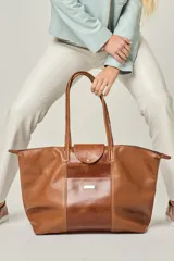 Bolso de viaje tipo tote de cuero color marrón, con doble asa de hombro, cierre superior con solapa y botón a presión, y bolsillo frontal con placa metálica.