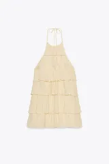Minivestido color crema con cuello halter ajustable con lazadas y espalda descubierta. Presenta volantes en el mismo tejido.
