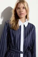 Vestido corto estilo camisero a rayas verticales azul marino y blancas, con cuello y puños blancos. Tiene cierre frontal con botones y lazo para atar en la cintura.