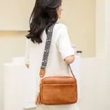 Cartera bandolera rectangular de eco cuero color marrón caramelo, con bolsillo frontal horizontal. La correa es ancha, de tela con estampado de leopardo en blanco y negro, y tiene detalles de cuero marrón en los enganches y un pequeño dije de borla colgante.