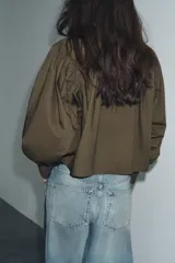 Camisa blanca de popelina con cuello solapa, manga larga abullonada con puño y bajo recto. Cierre frontal con botones ocultos por solapa.