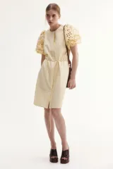Vestido color crema a la rodilla, de tejido liviano de algodón con cuello redondo, botones ocultos adelante y cinturón fino extraíble. Mangas al codo abullonadas en broderie con puños elásticos.