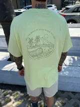 Remera color verde claro con estampado en la espalda de una ola, palmeras y montañas.