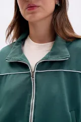 Campera deportiva de tejido de punto color verde oscuro, con cierre frontal plateado y cuello alto. Presenta vivos blancos que delinean el canesú y los puños y cintura son de elástico.
