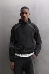Chaqueta deportiva negra con paneles grises en hombros y laterales, capucha ajustable, cierre frontal con cremallera, bolsillos laterales con cremallera y puños elásticos. Zara Athleticz.