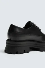 Zapato de vestir estilo derby color negro, con parte superior lisa, horma redonda y suela track con volumen.