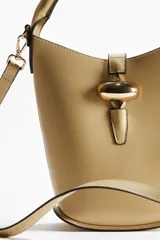Cartera tipo bucket color beige, con asa de mano tubular y correa cruzada ajustable y extraíble. Cierre con trabilla y adorno metálico dorado en el frente. Forrada.
