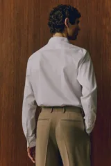 Camisa blanca de algodón con cuello italiano, tapeta lisa, canesú en la espalda y bajo redondeado. Mangas largas con tapeta de manga y botón, y cierre ajustable de botón en los puños. Corte ajustado que realza el contorno del cuerpo creando una silueta entallada.