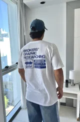 Remera blanca de manga corta con estampado gráfico en azul en la espalda, que incluye el texto "BROKERS GRAPHIC RESEARCHING" y varios logos de la marca.