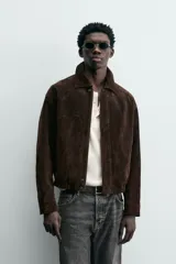 Cazadora de hombre de corte relajado confeccionada en piel con acabado ante, color marrón oscuro. Presenta cuello solapa, manga larga con puños abotonados a presión, bolsillos de vivo en cadera, bolsillo interior y bajo con elástico. Cierre frontal oculto por solapa con botones a presión.