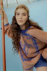 Buzo oversized de punto color tostado con diseño gráfico frontal en azul inspirado en una estampilla con una sirena y el texto 'Taormina'. Presenta tajos laterales, cuello redondo y terminaciones en contraste de color azul.