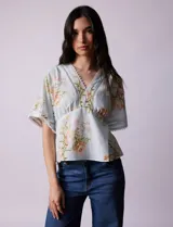 Blusa blanca estampada con flores azules, escote en V y mangas cortas amplias. Detalles de puntilla en escote y mangas.