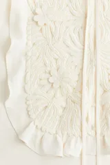 Blusa color crema de crepé de viscosa con mangas largas acampanadas y pechera con volados y bordado floral. Tiene cuello redondo y lazo para atar en el frente.