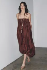 Vestido largo marrón de tejido satinado, con escote recto, tirantes finos extraíbles, bolsillos laterales ocultos y bajo asimétrico acabado en globo.