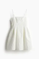 Vestido corto blanco en tejido jacquard con escote recto y breteles spaghetti ajustables. Modelo con cuerpo entallado, pliegues en la cintura y pollera con vuelo. Cierre oculto y cierre de corchete en la espalda. Parcialmente forrado.