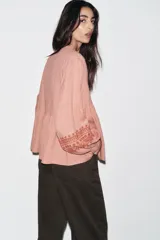 Blusa color rosa pálido de algodón con cuello redondo con volantes y cordón. Escote pico y manga larga. Detalle de bordados combinados a contraste color marrón. Cierre frontal con botones.