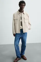 Sobrecamisa color beige de corte relaxed fit, confeccionada en tejido con elasticidad. Tiene cuello solapa, manga larga con puños abotonados, bolsillos de parche con solapa en el pecho y bolsillos de ojal en la cadera. El cierre es frontal con botones.