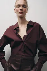 Camisa color borgoña de Zara Woman Collection, confeccionada en algodón popelín. Tiene cuello solapa, escote en pico, mangas largas con puños abotonados, bajo redondeado y cierre frontal con botones.