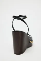 Sandalias negras con plataforma de madera, tacón de cuña y tira delantera con detalle de tachas doradas. Cierre mediante tira con pulsera al tobillo y acabado en punta cuadrada.