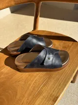 Sandalias de cuero color azul marino con plantilla dorada y suela marrón. Presentan una tira ancha cruzada con doble remache dorado.