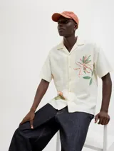 Camisa de manga corta color crema con estampado de flores y hojas en tonos naranja y verde. Tiene cuello resort, cierre frontal con botones y calce relajado.