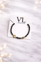 Pulsera elástica con cuentas facetadas de color azul oscuro iridiscente, separadas por cuentas metálicas doradas. Incluye dos dijes metálicos dorados: una cruz y una medalla religiosa.