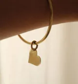 Brazalete rígido de metal color dorado con un dije colgante en forma de corazón liso.