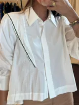 Camisa blanca de mangas largas con cuello clásico y cierre frontal con botones. Presenta un panel diagonal con detalle de ribete ondulado en verde. Corte recto.