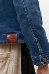 Chaqueta de jean azul medio, de corte clásico, con cuello camisero, bolsillos con solapa en el pecho y cierre frontal con botones metálicos color cobre.