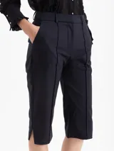 Pantalón capri estilo sastrero, color negro, de corte ajustado al cuerpo. Presenta alforzas en los delanteros, bolsillos ojales falsos y aberturas con tajo en los laterales inferiores.
