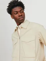Chaqueta color crema, de manga larga, con cuello camisero y cierre frontal con cremallera metálica. Tiene dos bolsillos de parche con solapa en el pecho.