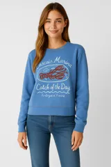 Buzo celeste de corte cropped, con cuello redondo y mangas largas. Estampado frontal con texto "Cascais Marina Catch of the Day Portuguese Riviera" y dibujo de una langosta.