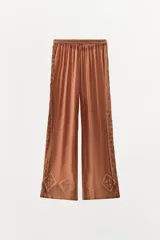Pantalón marrón de tiro alto con cintura elástica ajustable con lazada, corte palazzo y detalle de bordados en forma de rombo y puntilla a tono en el ruedo y laterales.