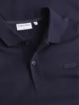 Chomba azul marino Calvin Klein de corte slim, confeccionada en algodón orgánico. Presenta cuello clásico con solapa de dos botones, mangas cortas con puños de punto y logo tonal bordado en el pecho.