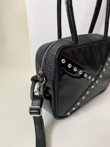Cartera rígida negra de cuero con tachas plateadas y tres combinaciones de cuero diferentes que aportan textura. Tiene herrajes y cierre metálico plateado marca YKK. Cuenta con un asa fina para llevar al hombro o en la mano y una correa larga extra regulable.