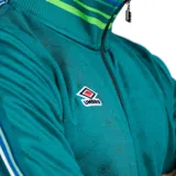 Campera deportiva Umbro color verde azulado con cuello alto, cierre frontal con cremallera, puños y cintura acanalados a rayas verdes, blancas y azules. Logo de Umbro bordado en el pecho.